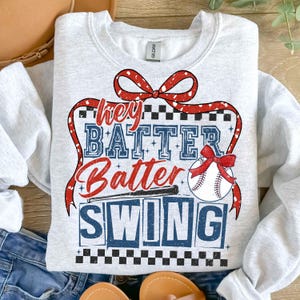 May include: A gray sweatshirt with a red, white, and blue baseball-themed graphic. The graphic says "Hey Batter Batter Swing" with a baseball and bat.