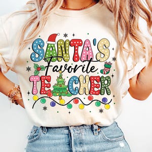 May include: A white t-shirt with a colorful graphic that says "Santa's Favorite Teacher" with a Christmas tree, lights, and a stocking.