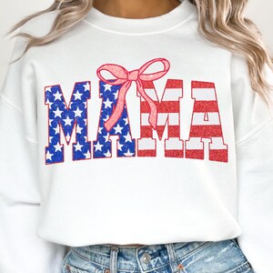 May include: White sweatshirt with a red, white, and blue "MAMA" graphic with a pink bow on top. The letters are in a glittery font.