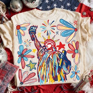 May include: Cream-colored t-shirt featuring a colorful illustration of the Statue of Liberty, surrounded by stylized flowers, stars, and fireworks. The design uses red, blue, yellow, and white colors, creating a patriotic theme.