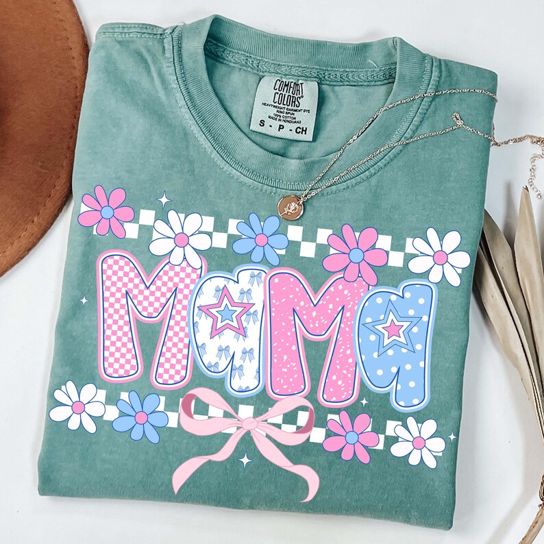 May include: A sage green t-shirt with the word "MAMA" in pink and blue, decorated with flowers and a pink bow. The shirt has a crew neck and short sleeves. A gold necklace with a pendant is draped across the shirt.