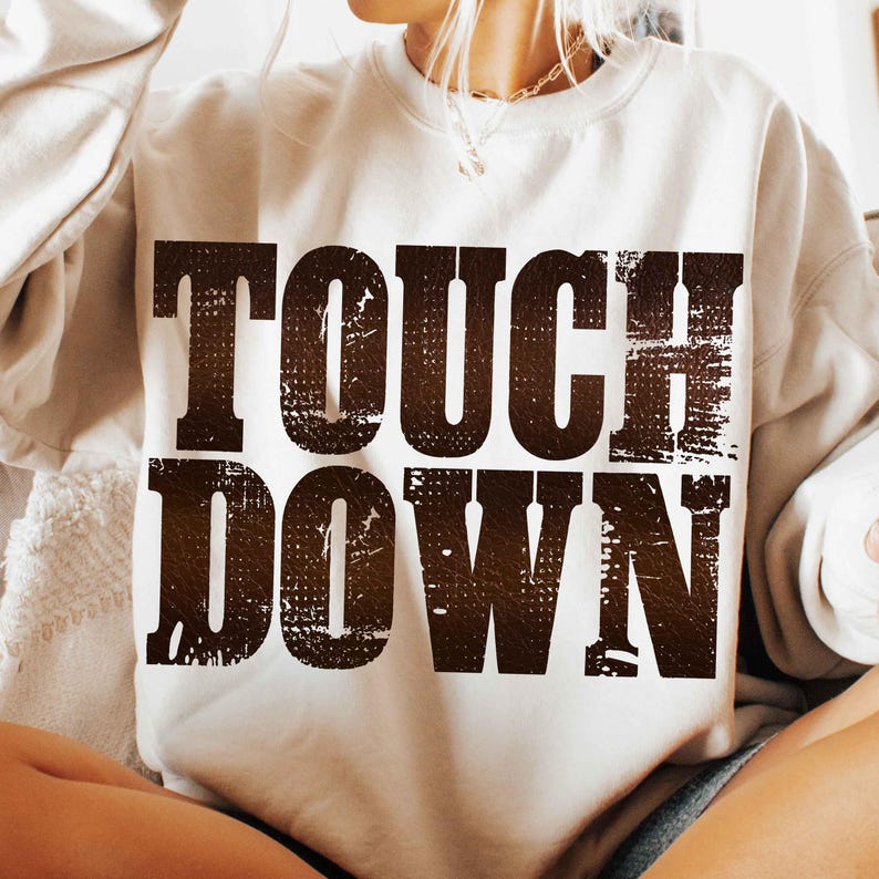 May include: A cream-colored sweatshirt with the words "TOUCH DOWN" printed in a distressed brown font. The sweatshirt is a casual style, suitable for everyday wear. The text is large and centered on the front.