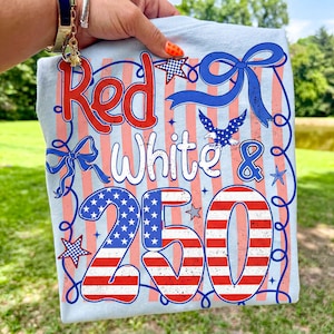 May include: Light blue t-shirt with a patriotic design. The shirt features the words "Red White & 250" in red and white, along with a blue ribbon, stars, and an eagle graphic. The design has a vintage, distressed look.