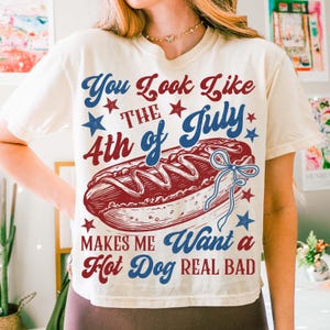 Puede incluir: Camiseta color crema con un dise&ntilde;o gr&aacute;fico que presenta un perrito caliente y texto. El texto dice "You Look Like The 4th of July Makes Me Want a Hot Dog REAL BAD" en rojo y azul. Estrellas dispersas alrededor del texto y el perrito caliente.