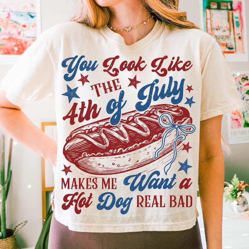 Puede incluir: Camiseta color crema con un dise&ntilde;o gr&aacute;fico que presenta un perrito caliente y texto. El texto dice "You Look Like The 4th of July Makes Me Want a Hot Dog REAL BAD" en rojo y azul. Estrellas dispersas alrededor del texto y el perrito caliente.