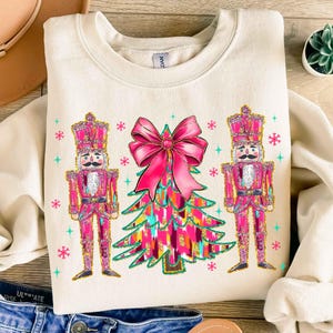 May include: A cream-colored sweatshirt with a pink and gold Christmas tree design. The tree is decorated with a large pink bow and two pink and gold nutcracker figures.