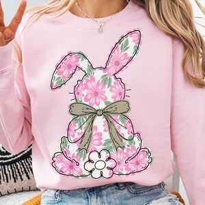 May include: Pink sweatshirt with a floral bunny design. The bunny is outlined in black and has a brown bow. The bunny is surrounded by pink flowers.