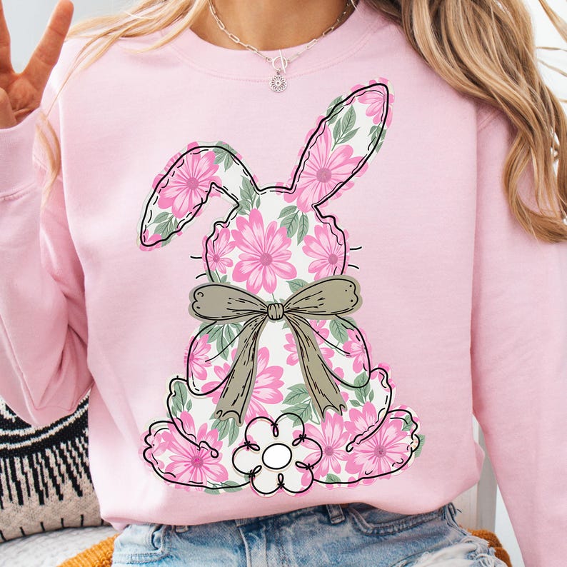May include: Pink sweatshirt with a floral bunny design. The bunny is outlined in black and has a brown bow. The bunny is surrounded by pink flowers.