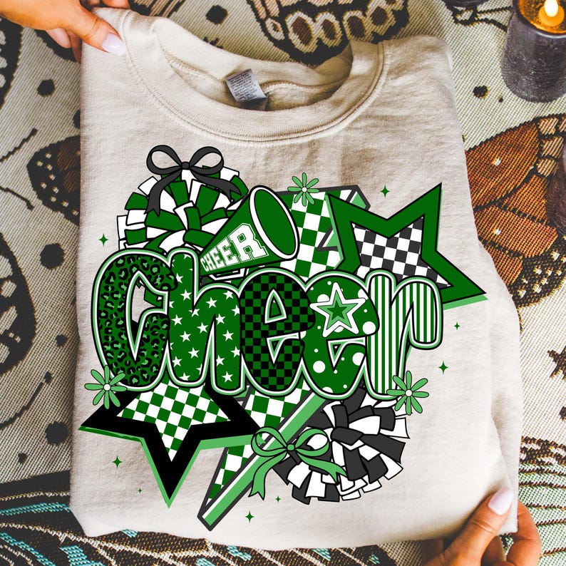 May include: A beige sweatshirt with a green and black "CHEER" graphic. The design includes stars, pom-poms, and a megaphone. The word "CHEER" is in various fonts and patterns, including leopard print and checkered designs.