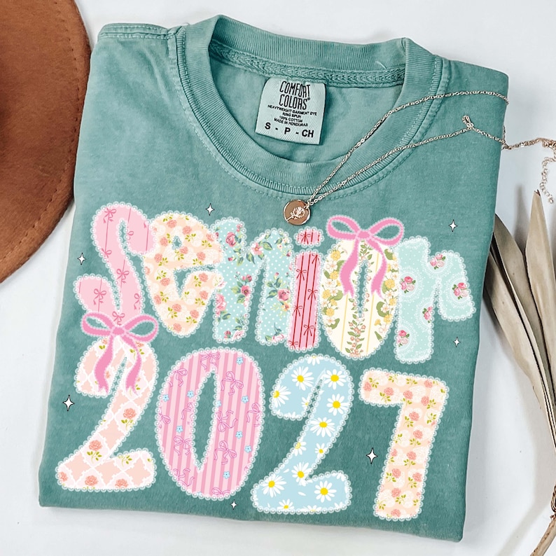 May include: A teal Comfort Colors t-shirt with the words "Senior 2027" in a patchwork design. The letters and numbers are decorated with floral patterns, bows, and daisies. A silver necklace with a pendant rests on the shirt.