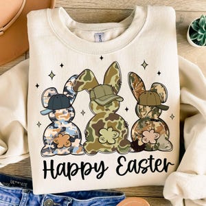 May include: A beige sweatshirt with a graphic of three camouflage Easter bunnies wearing baseball caps. The bunnies are standing in a row and the text "Happy Easter" is printed below them.