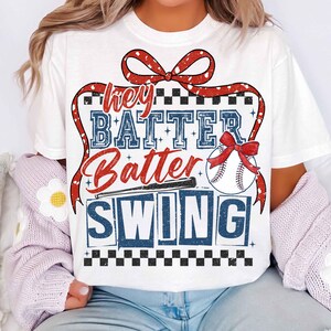 May include: White t-shirt with a red, white, and blue baseball-themed graphic. The graphic features a red bow, a baseball, a bat, and the text "Hey Batter Batter Swing".
