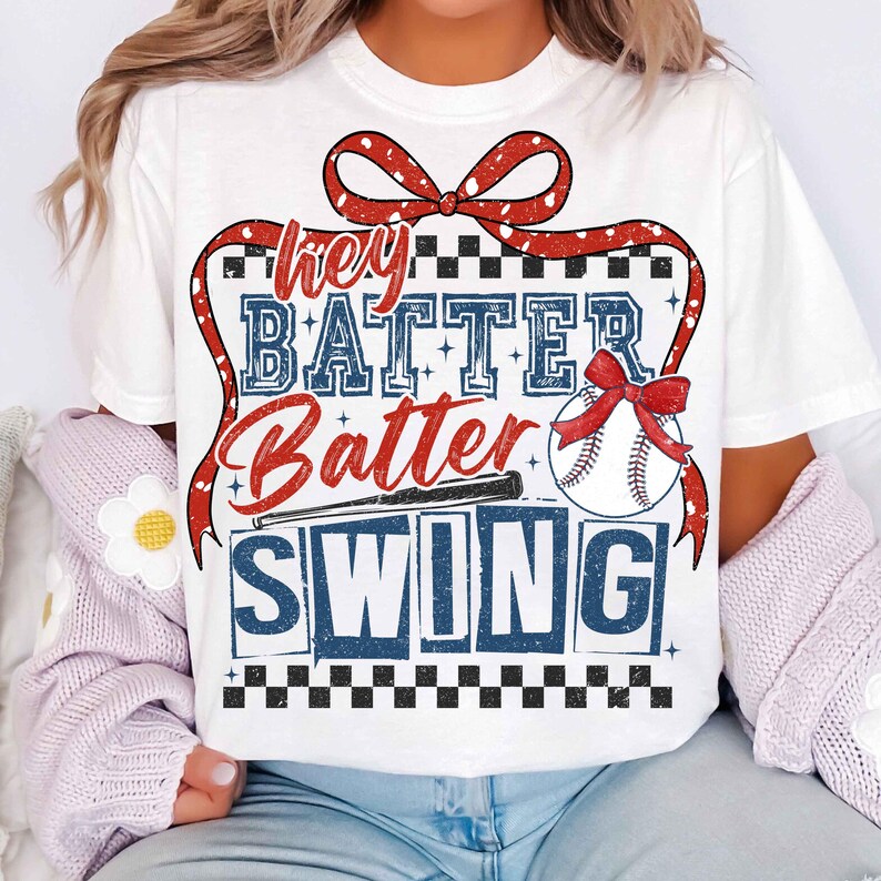 May include: White t-shirt with a red, white, and blue baseball-themed graphic. The graphic features a red bow, a baseball, a bat, and the text "Hey Batter Batter Swing".