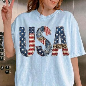 Pu&ograve; includere: T-shirt azzurra con la scritta "USA" in un design patchwork. Le lettere presentano denim, stampa leopardata e la bandiera americana. La maglietta ha maniche corte e girocollo.
