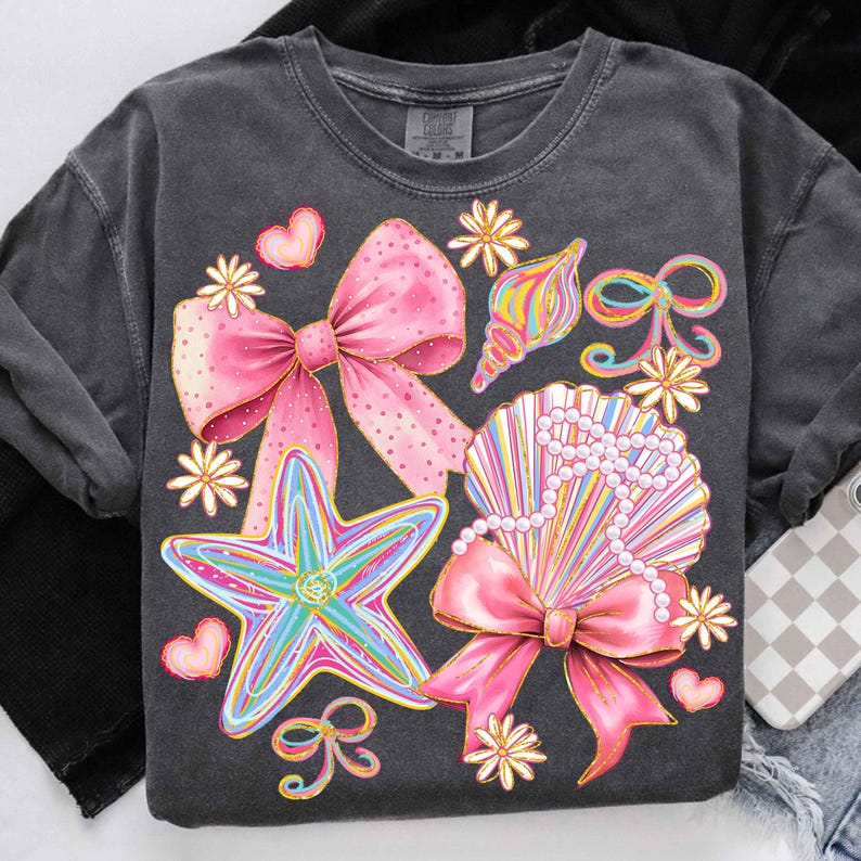 Op de afbeelding: Donkergrijs T-shirt met een kleurrijk grafisch ontwerp. Het ontwerp bevat een roze strik, zeesterren, schelpen en bloemen. De zeester is blauw, groen en geel. De strik is roze met gouden accenten.