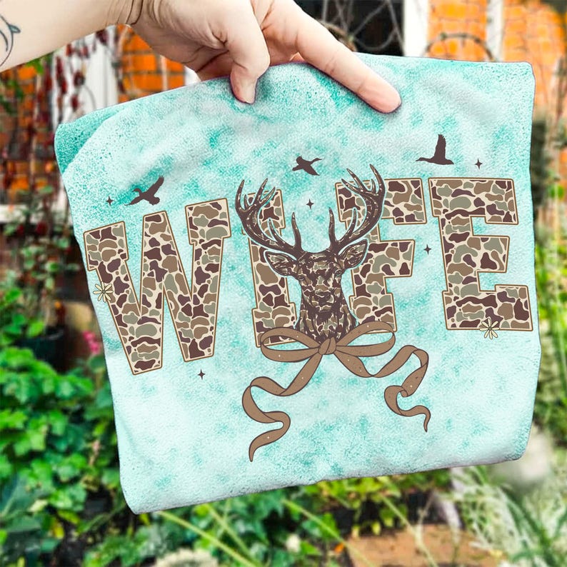 May include: A teal-colored graphic tee featuring the word "WIFE" in camouflage print. A deer head with antlers and a brown bow is centered below the word. The design includes flying birds and small stars.