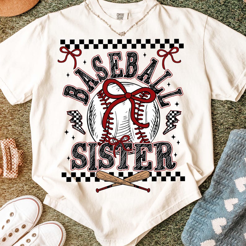 May include: Cream-colored t-shirt with a baseball-themed graphic. The design features a baseball with a red bow, the words "BASEBALL SISTER," and crossed baseball bats. Accents include black and white checkered patterns and lightning bolts.