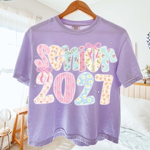 May include: A lilac-colored t-shirt with the word "Senior" and the number "2021" in a patchwork design. The letters and numbers feature floral and patterned fabrics, with pink bows and star accents.