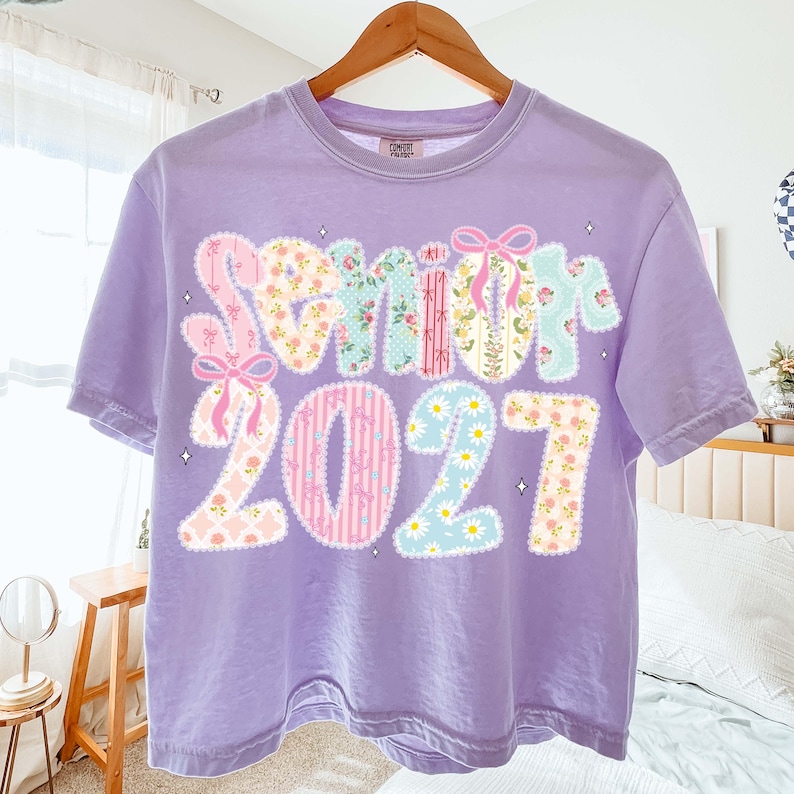 May include: A lilac-colored t-shirt with the word "Senior" and the number "2021" in a patchwork design. The letters and numbers feature floral and patterned fabrics, with pink bows and star accents.