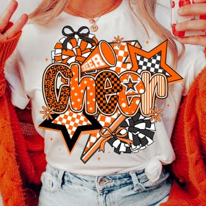 May include: White t-shirt with a large orange and black "Cheer" graphic. The design includes stars, pom-poms, and a megaphone. The word "Cheer" is in a mix of fonts and patterns, including leopard print and checkered designs.