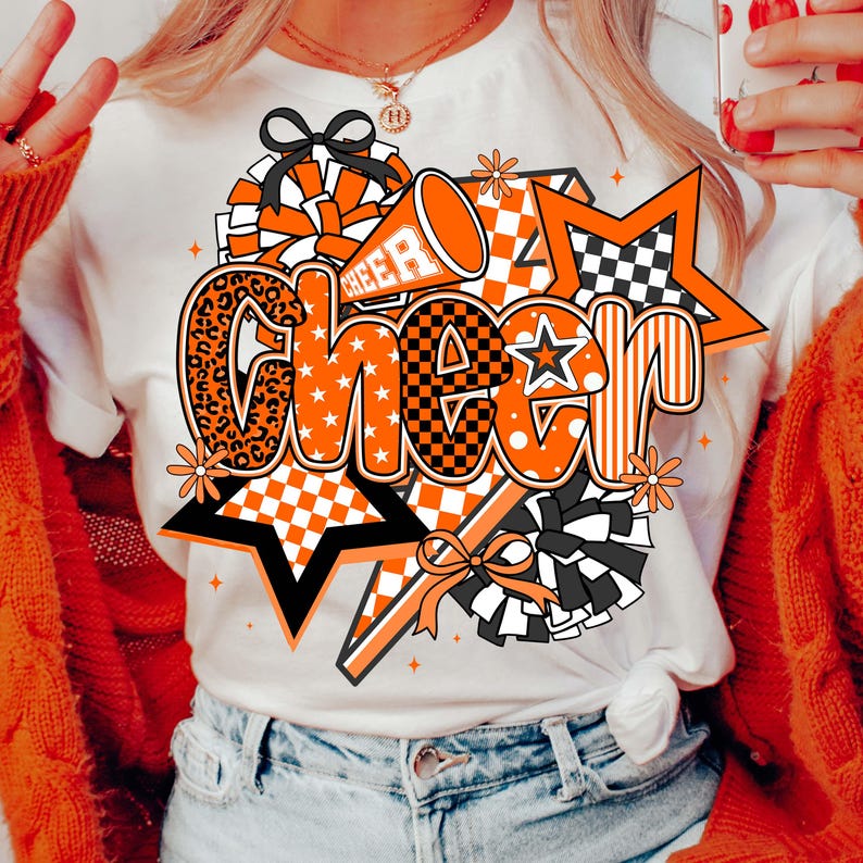 May include: White t-shirt with a large orange and black "Cheer" graphic. The design includes stars, pom-poms, and a megaphone. The word "Cheer" is in a mix of fonts and patterns, including leopard print and checkered designs.