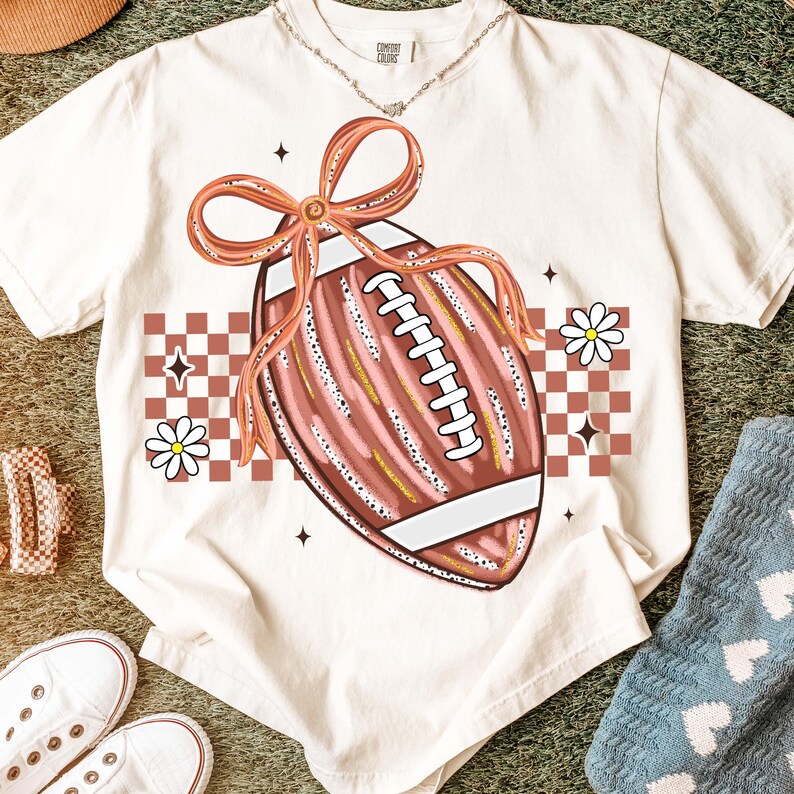 May include: Cream-colored t-shirt featuring a football graphic with a bow, daisies, and a checkered pattern. The football is brown and pink with white laces. The shirt has a relaxed fit and short sleeves.
