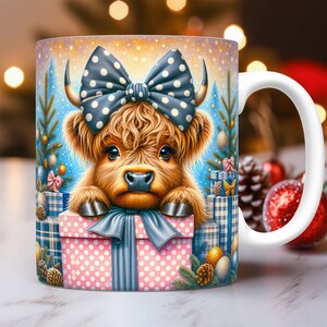 May include: A white ceramic mug with a festive design featuring a brown and white Highland cow wearing a blue and white polka dot bow. The cow is peeking out of a pink and white polka dot gift box with a blue ribbon. The mug is surrounded by Christmas decorations, including a small pine tree, ornaments, and a red and white striped gift box.