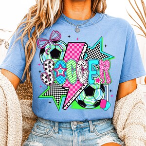 Puede incluir: Una camiseta azul claro con un dise&ntilde;o gr&aacute;fico de la palabra "SOCCER" en letras de colores con un rayo, estrellas y balones de f&uacute;tbol.