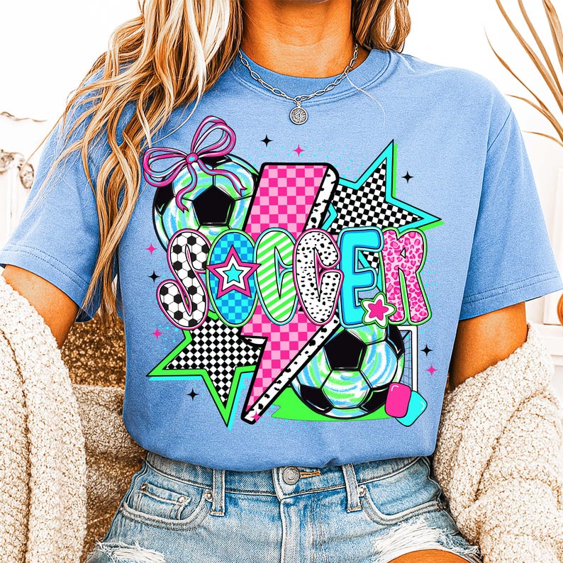 Puede incluir: Una camiseta azul claro con un dise&ntilde;o gr&aacute;fico de la palabra "SOCCER" en letras de colores con un rayo, estrellas y balones de f&uacute;tbol.