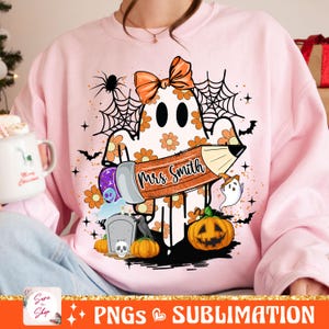 Puede incluir: Sudadera rosa con un fantasma sosteniendo un l&aacute;piz que dice "Mrs. Smith" con un lazo en la cabeza. El fantasma est&aacute; rodeado de calabazas, murci&eacute;lagos y telara&ntilde;as.