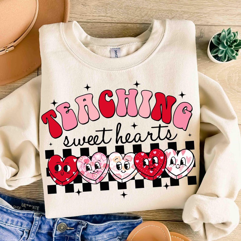 Pu&ograve; includere: Una felpa beige con il testo "TEACHING sweet hearts" in lettere rosa e rosse. Il testo &egrave; sopra una fila di cinque cuori di cartoni animati con facce su uno sfondo a scacchi bianco e nero.