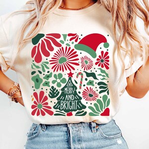 Puede incluir: Sudadera de color crema con un dise&ntilde;o navide&ntilde;o festivo. El gr&aacute;fico incluye elementos florales rojos y verdes, un gorro de Pap&aacute; Noel, un bast&oacute;n de caramelo y un &aacute;rbol de Navidad con las palabras "MERRY AND BRIGHT".