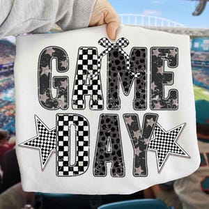 May include: White fabric with the words "GAME DAY" in a distressed, black and gray design. The letters have a mix of patterns, including stars, polka dots, and checkered flags. Two star shapes with checkered patterns flank the word "DAY".