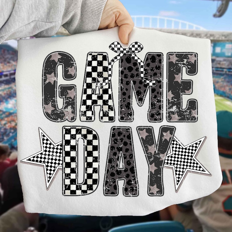 May include: White fabric with the words "GAME DAY" in a distressed, black and gray design. The letters have a mix of patterns, including stars, polka dots, and checkered flags. Two star shapes with checkered patterns flank the word "DAY".