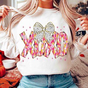 K&ouml;nnte beinhalten: Wei&szlig;es Sweatshirt mit den Buchstaben XOXO in rosa, gelben und wei&szlig;en Mustern und einer schwarz-wei&szlig; gepunkteten Schleife. Rosa Sternakzente umgeben die Buchstaben. Ein l&auml;ssiger, bequemer Stil.