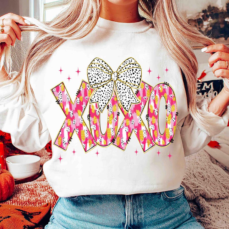 K&ouml;nnte beinhalten: Wei&szlig;es Sweatshirt mit den Buchstaben XOXO in rosa, gelben und wei&szlig;en Mustern und einer schwarz-wei&szlig; gepunkteten Schleife. Rosa Sternakzente umgeben die Buchstaben. Ein l&auml;ssiger, bequemer Stil.