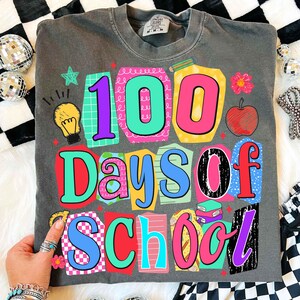 Puede incluir: Camiseta gris oscuro con un gr&aacute;fico colorido que celebra "100 d&iacute;as de escuela". El dise&ntilde;o presenta los n&uacute;meros "100" en colores brillantes, junto con las palabras "Days of School". Elementos adicionales incluyen una manzana, libros y una bombilla.
