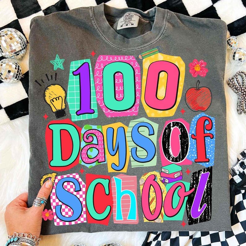 Puede incluir: Camiseta gris oscuro con un gr&aacute;fico colorido que celebra "100 d&iacute;as de escuela". El dise&ntilde;o presenta los n&uacute;meros "100" en colores brillantes, junto con las palabras "Days of School". Elementos adicionales incluyen una manzana, libros y una bombilla.