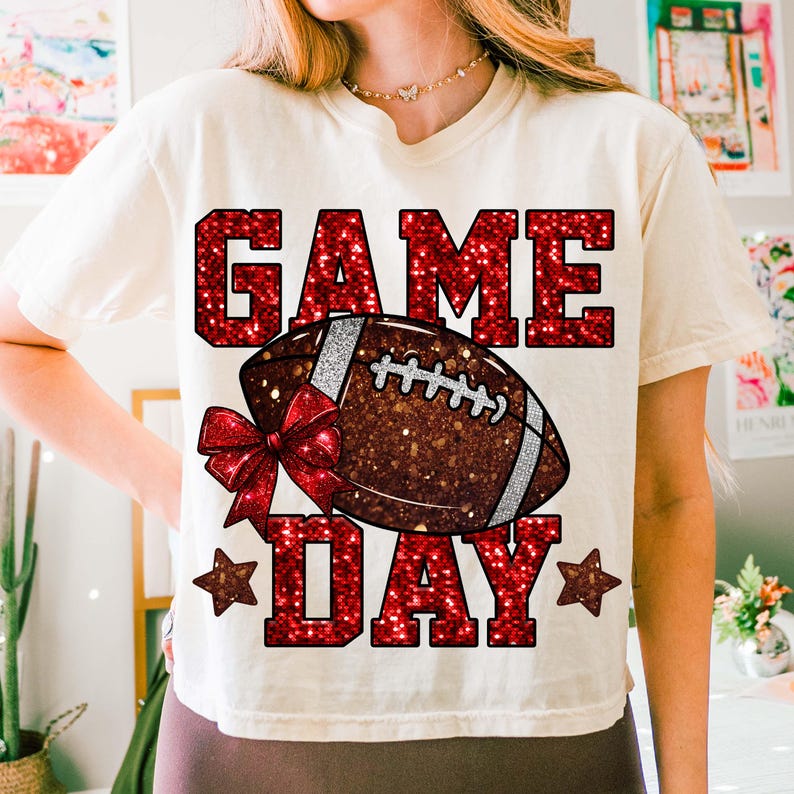 Puede incluir: Camiseta color crema con un dise&ntilde;o rojo brillante de "GAME DAY". El dise&ntilde;o presenta un bal&oacute;n de f&uacute;tbol con rayas blancas y un lazo rojo. Dos estrellas rojas flanquean la palabra "DAY".