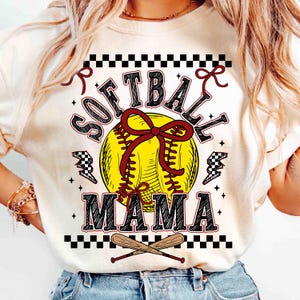 May include: Cream-colored t-shirt with a softball graphic. The design features a yellow softball with a red bow, the words "SOFTBALL MAMA" in black and red, and crossed baseball bats at the bottom. Black and white checkered accents.