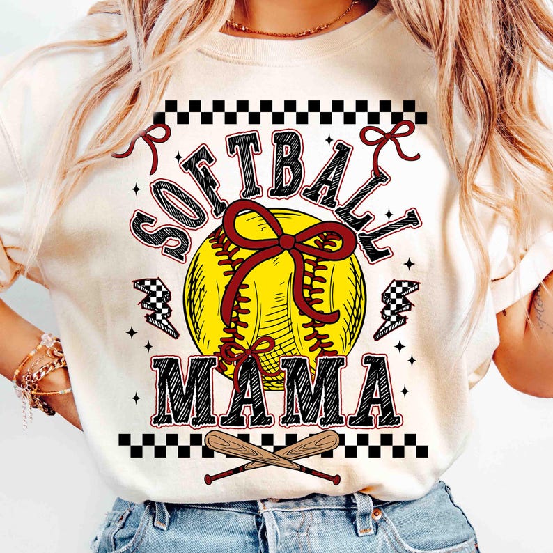 May include: Cream-colored t-shirt with a softball graphic. The design features a yellow softball with a red bow, the words "SOFTBALL MAMA" in black and red, and crossed baseball bats at the bottom. Black and white checkered accents.