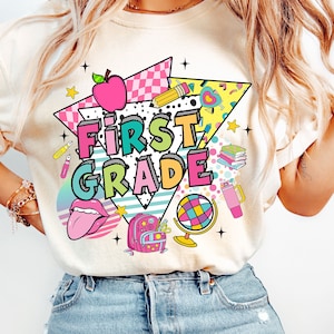 K&ouml;nnte beinhalten: Ein wei&szlig;es T-Shirt mit einem bunten Grafikdesign, auf dem "First Grade" steht. Das Design enth&auml;lt einen Regenbogen, einen Globus, einen Rucksack, einen Bleistift, ein Herz und einen Stern.