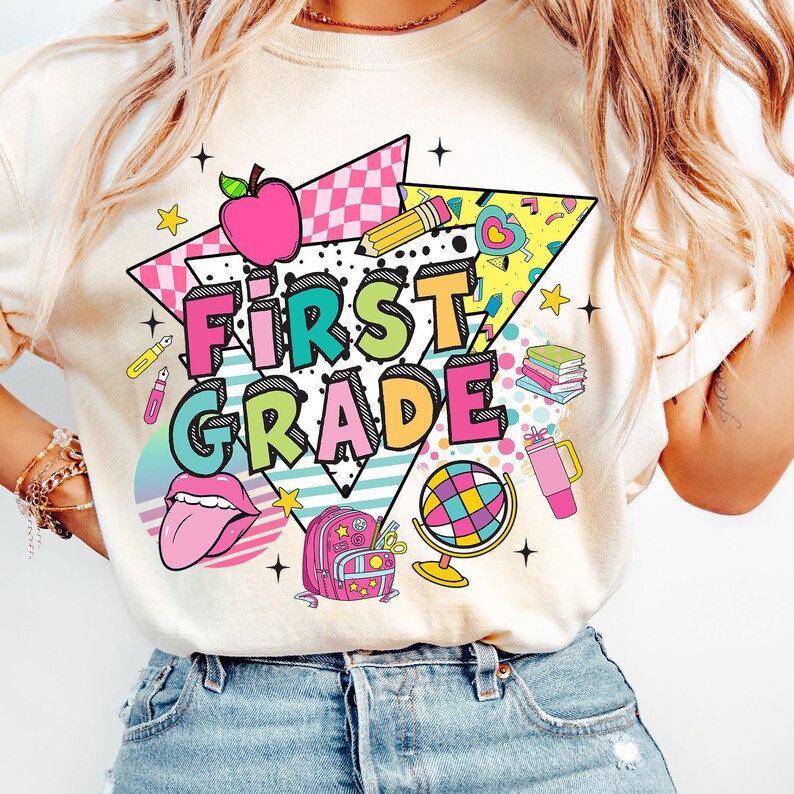 K&ouml;nnte beinhalten: Ein wei&szlig;es T-Shirt mit einem bunten Grafikdesign, auf dem "First Grade" steht. Das Design enth&auml;lt einen Regenbogen, einen Globus, einen Rucksack, einen Bleistift, ein Herz und einen Stern.