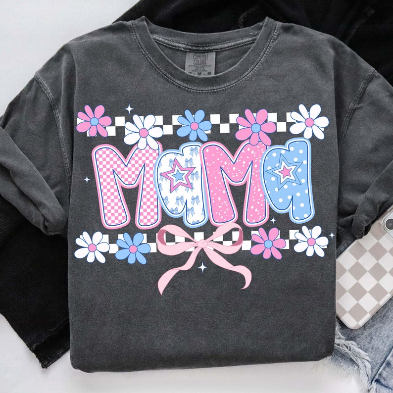May include: Dark gray sweatshirt with the word "Mama" in pink and blue bubble letters. The letters have a checkered pattern and star accents. The design includes a pink bow and floral elements in pink, white, and blue.