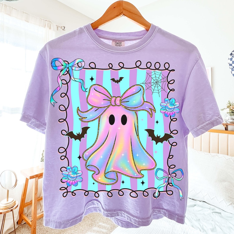 Puede incluir: Camiseta lila con un dise&ntilde;o de fantasma de dibujos animados, con un gran lazo, murci&eacute;lagos y flores. El fantasma est&aacute; enmarcado sobre un fondo a rayas. La camiseta est&aacute; colgada de una percha de madera.