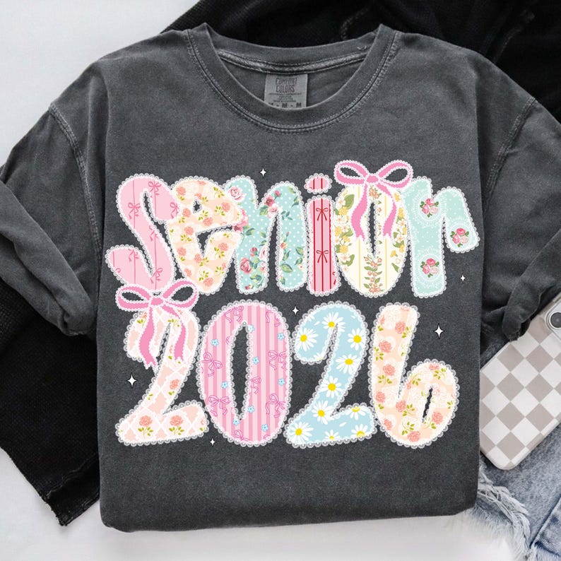 Op de afbeelding: Donkergrijze sweatshirt met de tekst "Senior 2026" in een kleurrijk patchwork-ontwerp. De letters en cijfers zijn versierd met bloempatronen, strepen en strikken, met een geschulpte rand.