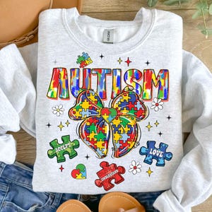 May include: A gray sweatshirt with a colorful tie-dye design that says "AUTISM" with a puzzle piece bow and the words "ACCEPT", "UNDERSTAND", and "LOVE" on puzzle pieces.