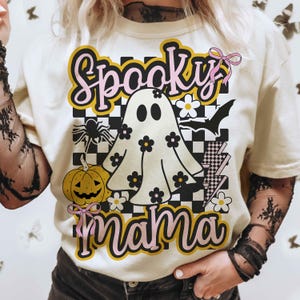 Puede incluir: Camiseta color crema con un dise&ntilde;o de Halloween. El dise&ntilde;o presenta las palabras "Spooky Mama" en rosa y amarillo, un fantasma con flores, una calabaza, una ara&ntilde;a y un murci&eacute;lago.