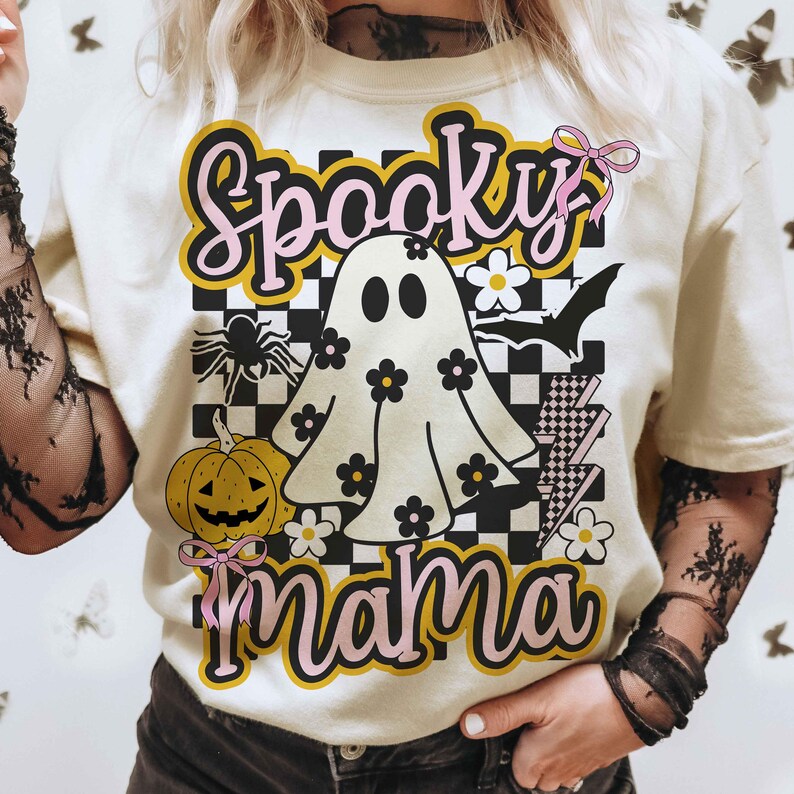 Puede incluir: Camiseta color crema con un dise&ntilde;o de Halloween. El dise&ntilde;o presenta las palabras "Spooky Mama" en rosa y amarillo, un fantasma con flores, una calabaza, una ara&ntilde;a y un murci&eacute;lago.