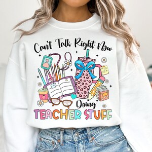 May include: White sweatshirt with a graphic design of school supplies and the text "Can't Talk Right Now Doing Teacher Stuff".